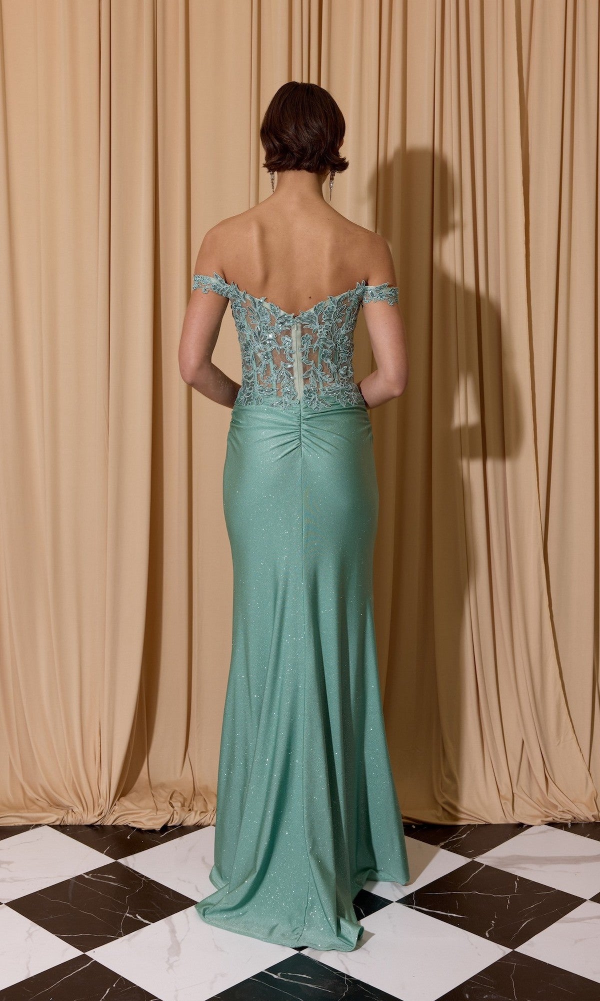 A person wearing the Long Prom Dress: Eureka EK173 in mint green, off-the-shoulder style stands with their back to the camera before beige curtains on a black and white tiled floor.
